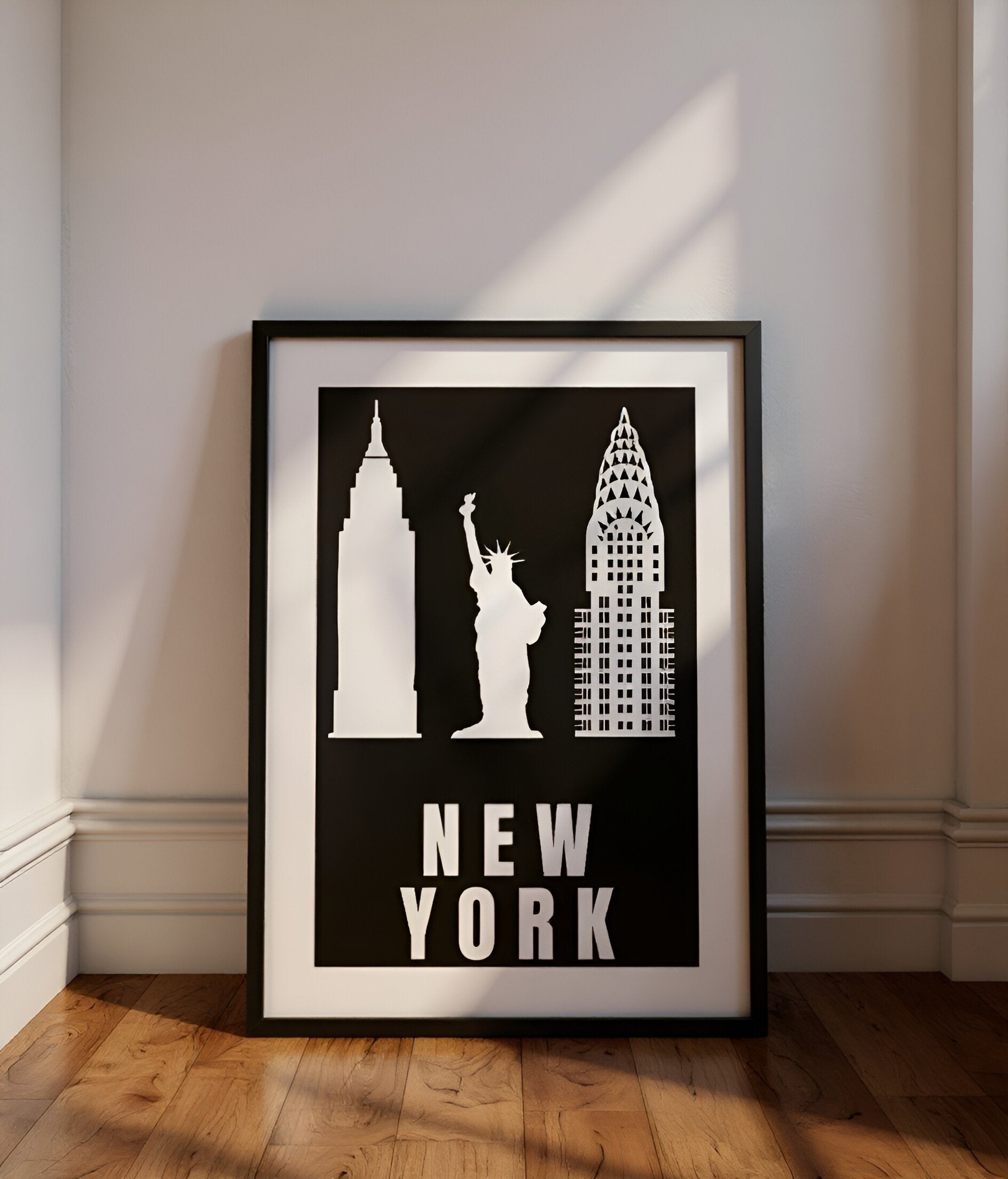 Framed poster of New York cityscape with Empire State Building, Statue of Liberty, and Chrysler Building on a black background. leaning against a cream wall with wooden floorboards and ambient sunlight shining through window.